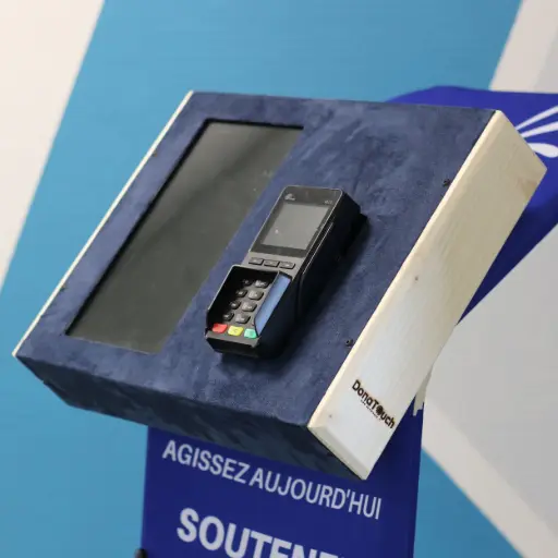 Unité de don mobile DonaTouch. Écran tactile et terminal TPE pour le paiement sécurisé sans contact et par carte.  Exporter vers Sheets Gros plan sur un pupitre de don DonaTouch (ou iTouchAm) avec un revêtement en suédine bleue et un panneau latéral en bois clair, montrant un terminal de paiement électronique prêt à l'emploi.