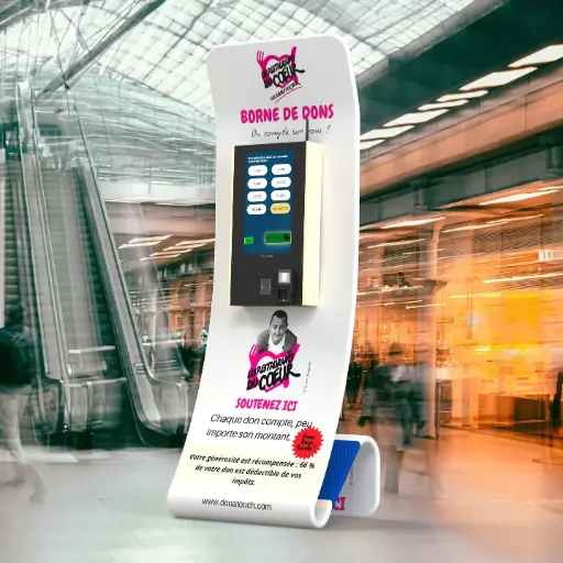 Borne de dons numérique des Restos du Cœur. Facilite le don par carte pour financer les repas et met en avant la déductibilité fiscale. Borne de dons DonaTouch aux couleurs des Restos du Cœur (blanc et rouge), affichant le message "BORNE DE DONS - On compte sur vous !" dans un hall de gare ou d'aéroport.